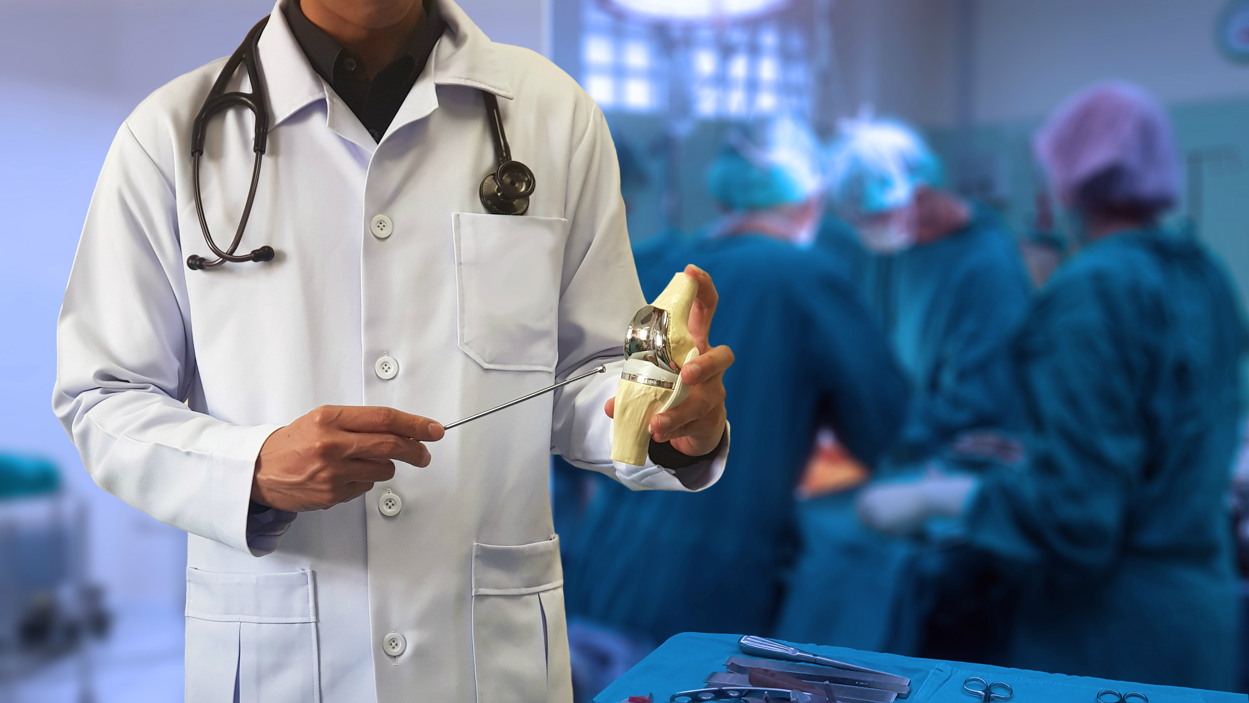 Orthopedic doctor showing model of Total knee replacement with blurred background of surgical team performing surgery in operating room. Medical technology concept.